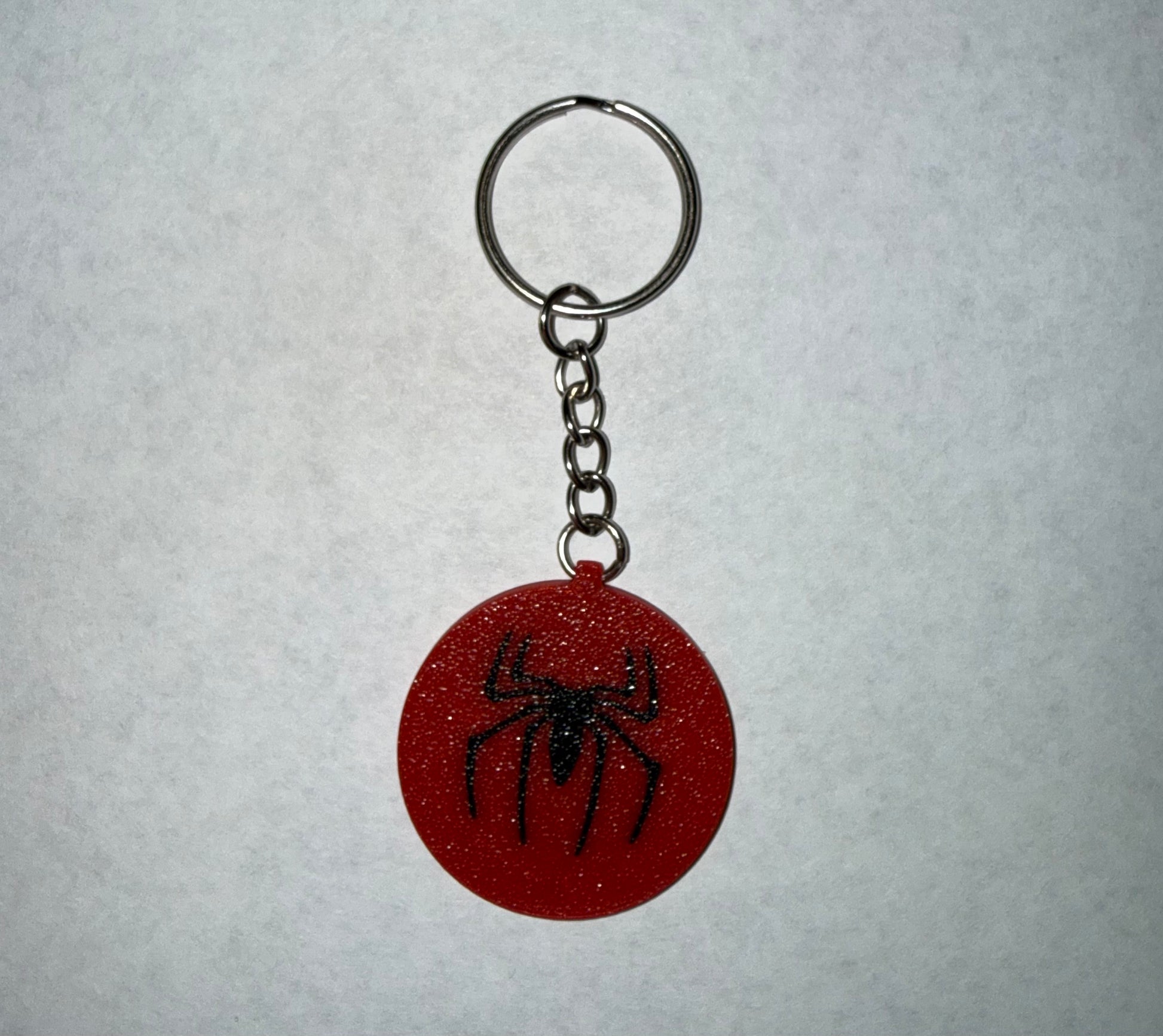 Red comic-inspired spider emblem keychain with black spider logo attached to a metal key ring on a white background.